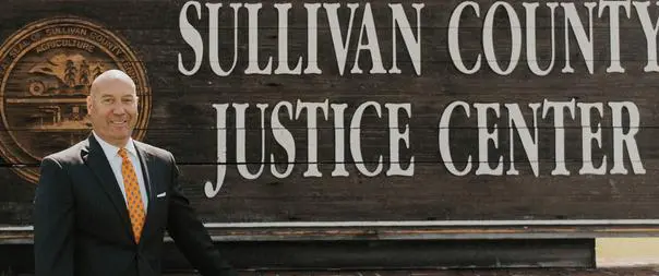 Man in suit standing by Sullivan County Justice Center sign.